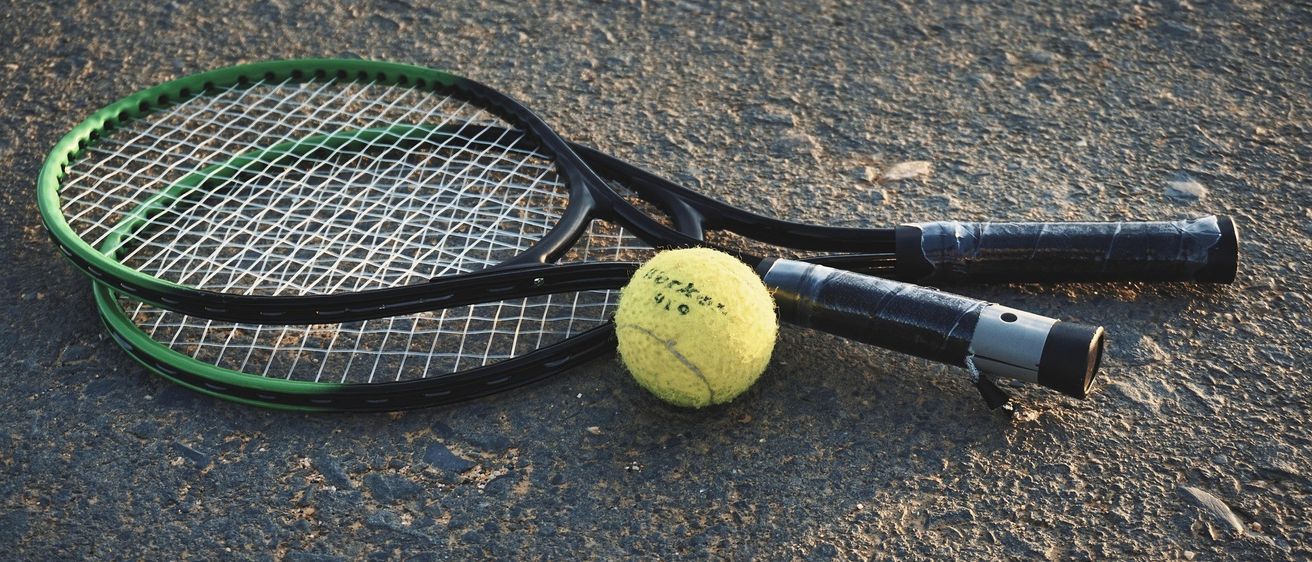 Ein Tennisball und zwei Tennisschläger liegen auf dem Boden. Die Schläger sind schwarz mit grünen Akzenten. Der Ball ist gelb mit schwarzen Schriftzügen.