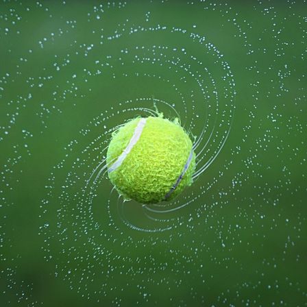Ein Tennisball in der Luft, umgeben von Wassertropfen, die einen dynamischen und lebendigen Effekt erzeugen.