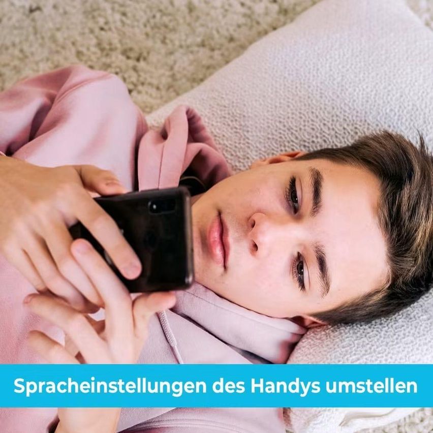 A young man lies on a bed, holding a smartphone, and appears to be reading or texting. He is wearing a pink hoodie.