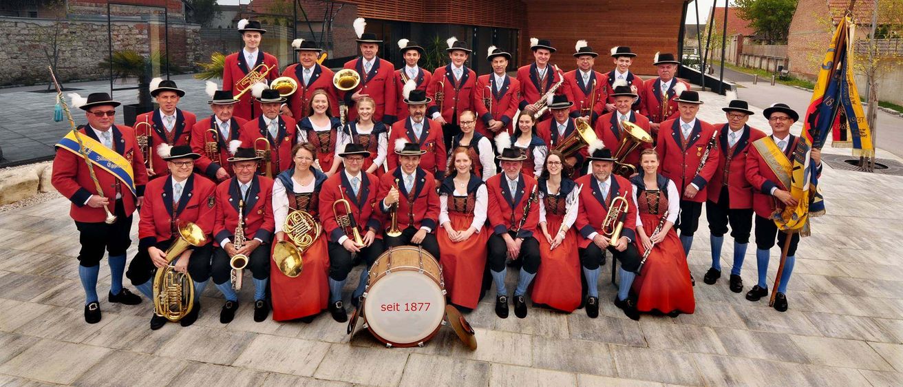 Eine Gruppe von Musikern in roten Uniformen posiert mit ihren Instrumenten vor einem Gebäude. Auf einem Trommel in der Mitte steht das Datum 'seit 1877'.