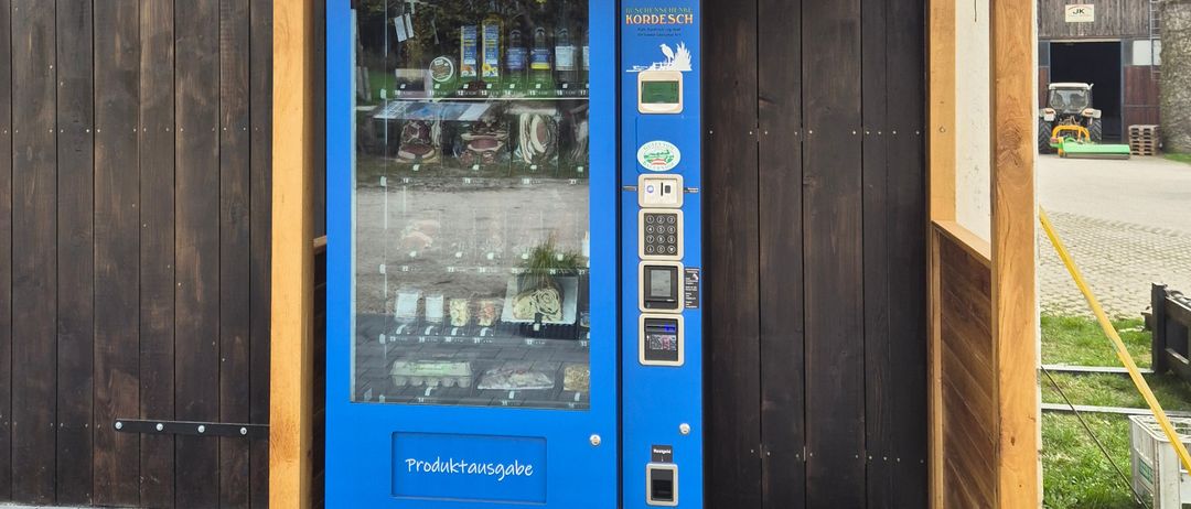 A blue vending machine under a wooden canopy. The machine has glass doors showing products inside. The word Produktauswahl is printed on it.