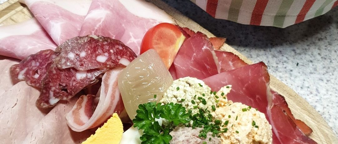Ein Holzteller mit verschiedenen Fleischsorten wie geschnittener Salami, Prosciutto und Mortadella, zusammen mit einem gekochten Ei, Gurken und Käse. Ein Korb mit Brot ist im Hintergrund.