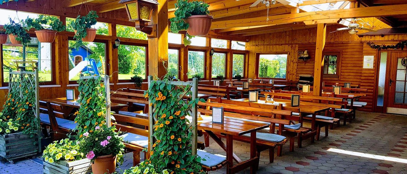 Ein Essbereich im Freien mit Holztischen und -bänken unter einem Holzdach mit Oberlichtern und Deckenventilatoren, dekoriert mit hängenden Pflanzen.