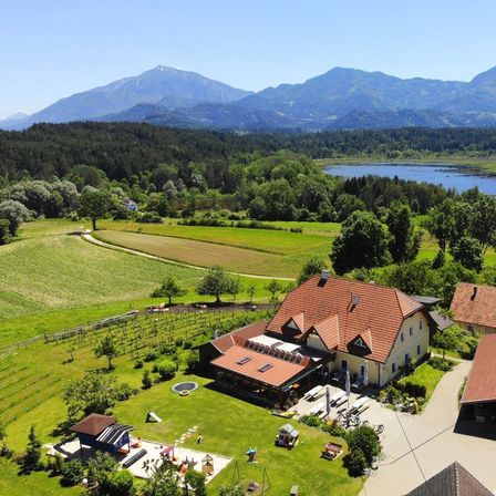 Ein Luftbild eines großen gelben Hauses mit einem braunen Dach, umgeben von grünen Feldern und einem See mit Bergen im Hintergrund.