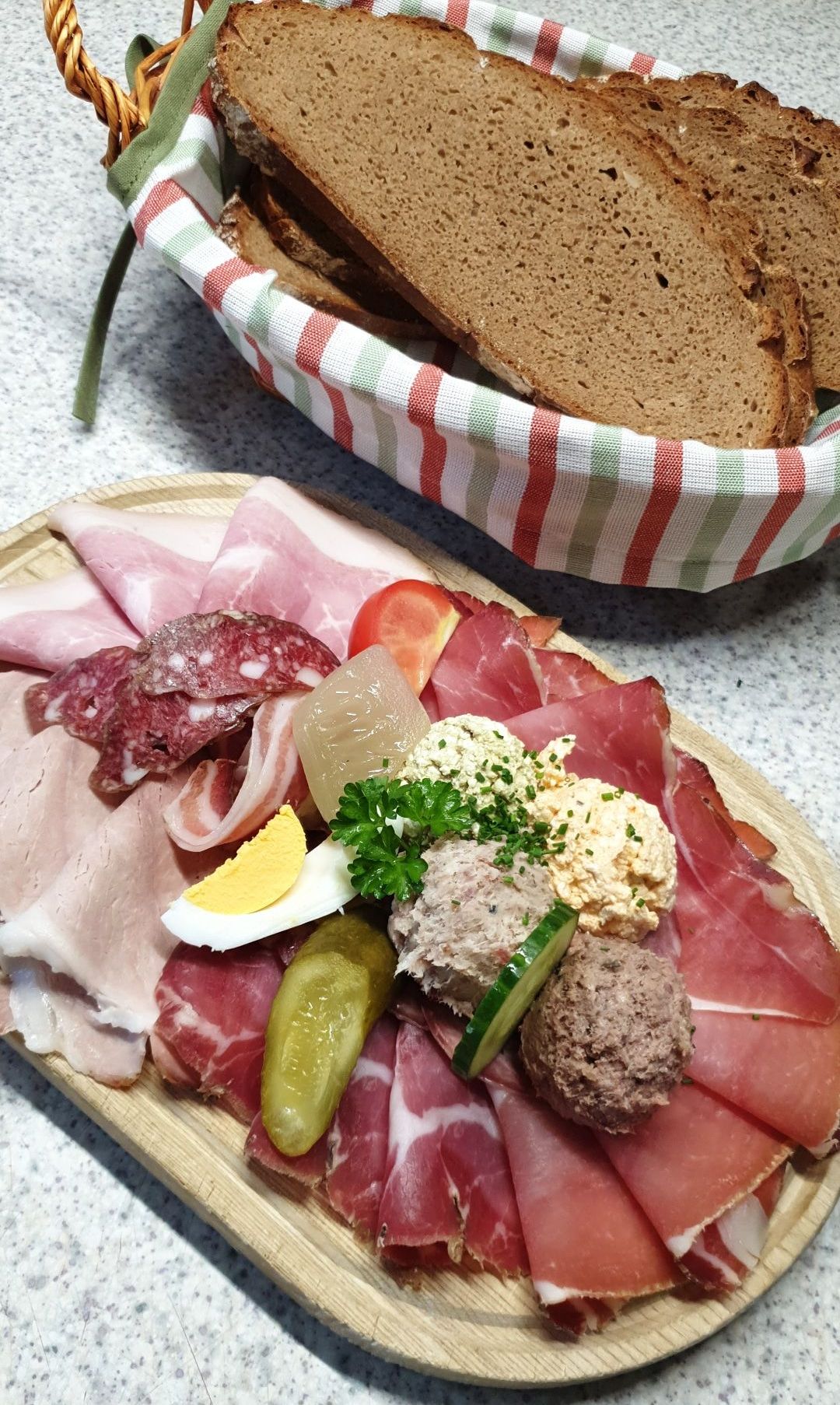Ein Holzbrett mit verschiedenen Fleischsorten, darunter Salami und Prosciutto, und Beilagen wie Eier und Gurken. Im Hintergrund befindet sich ein Brotkorb.