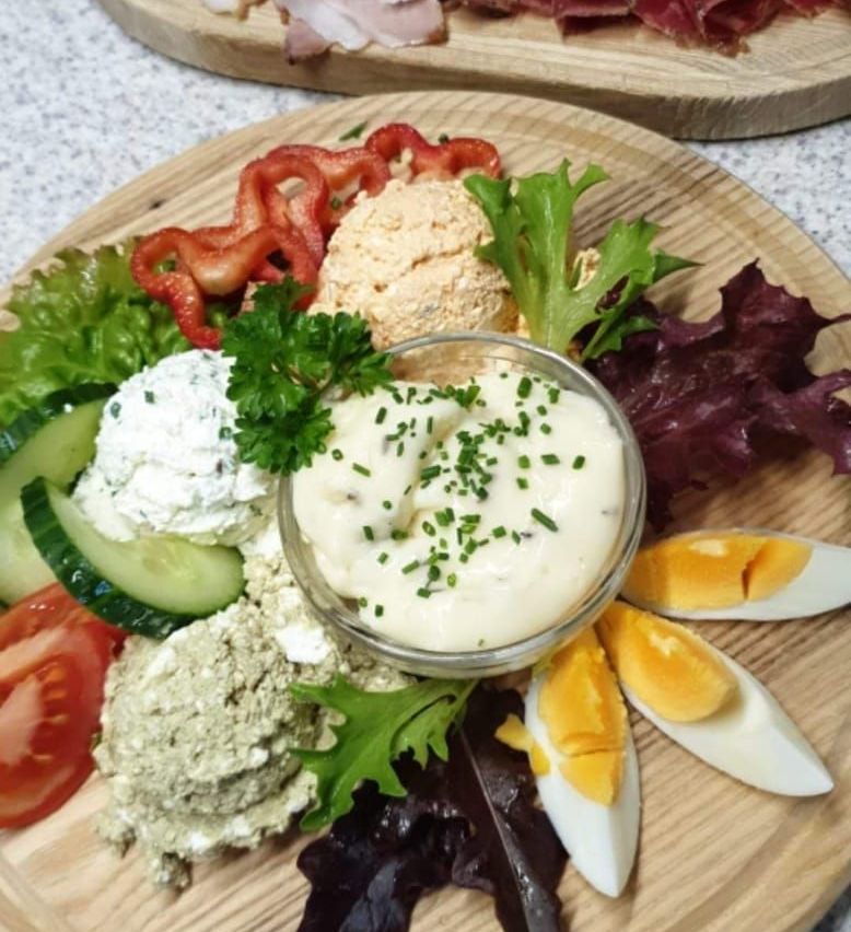 Ein Holztablett hält verschiedene Lebensmittel wie Hummus, Tzatziki, Eier, Gurken, Salat und Paprika. Das Gericht ist mit Petersilie und Schnittlauch garniert.