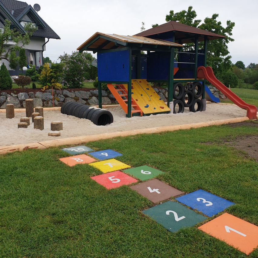 Ein farbenfroher Spielplatz mit Rutsche, Kletterwand und nummerierten Fliesen auf dem Rasen. Hölzerne Stämme umgeben einen Sandkasten. Bäume und ein Haus im Hintergrund.