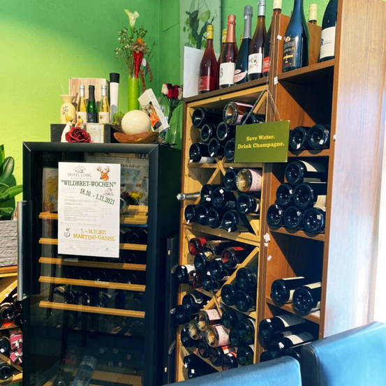 Ein Weinladen mit grüner Wand zeigt Flaschen Wein, einen Weinkühler mit einem Schild 'Wildbret-Wochen' und ein Schild 'Sparen Sie Wasser. Trinken Sie Champagner.'