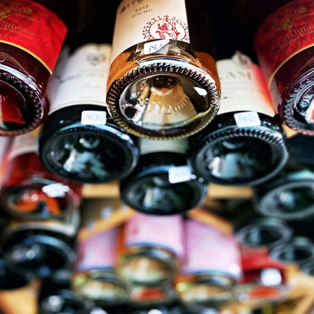 Mehrere Weinflaschen sind aufeinandergestapelt, mit einer klaren Glasflasche in der Mitte. Jede Flasche hat ein anderes Etikettendesign und eine andere Farbe.