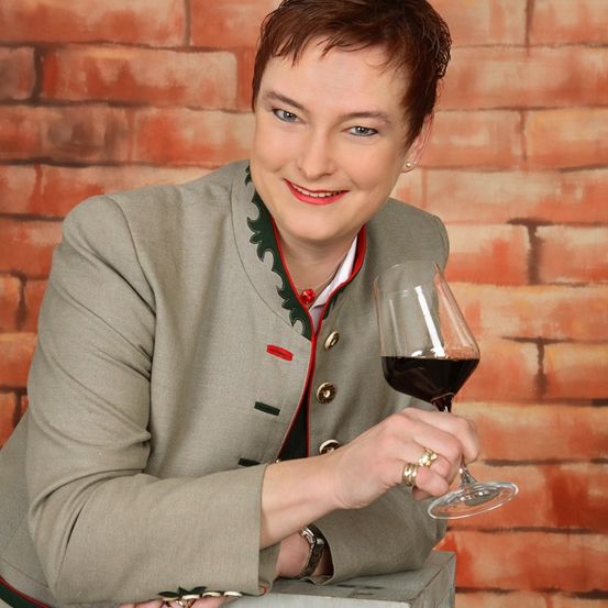 Eine Frau in einer grauen Jacke mit rotem Rand lächelt, während sie ein Glas Rotwein in der Hand hält. Hinter ihr ist eine Ziegelmauer.