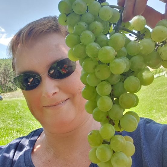 Eine Frau lächelt und hält eine Traube grüner Trauben in der Hand. Sie trägt eine Sonnenbrille und ein blaues Shirt. Hinter ihr ist ein Grasfeld und ein klarer blauer Himmel.