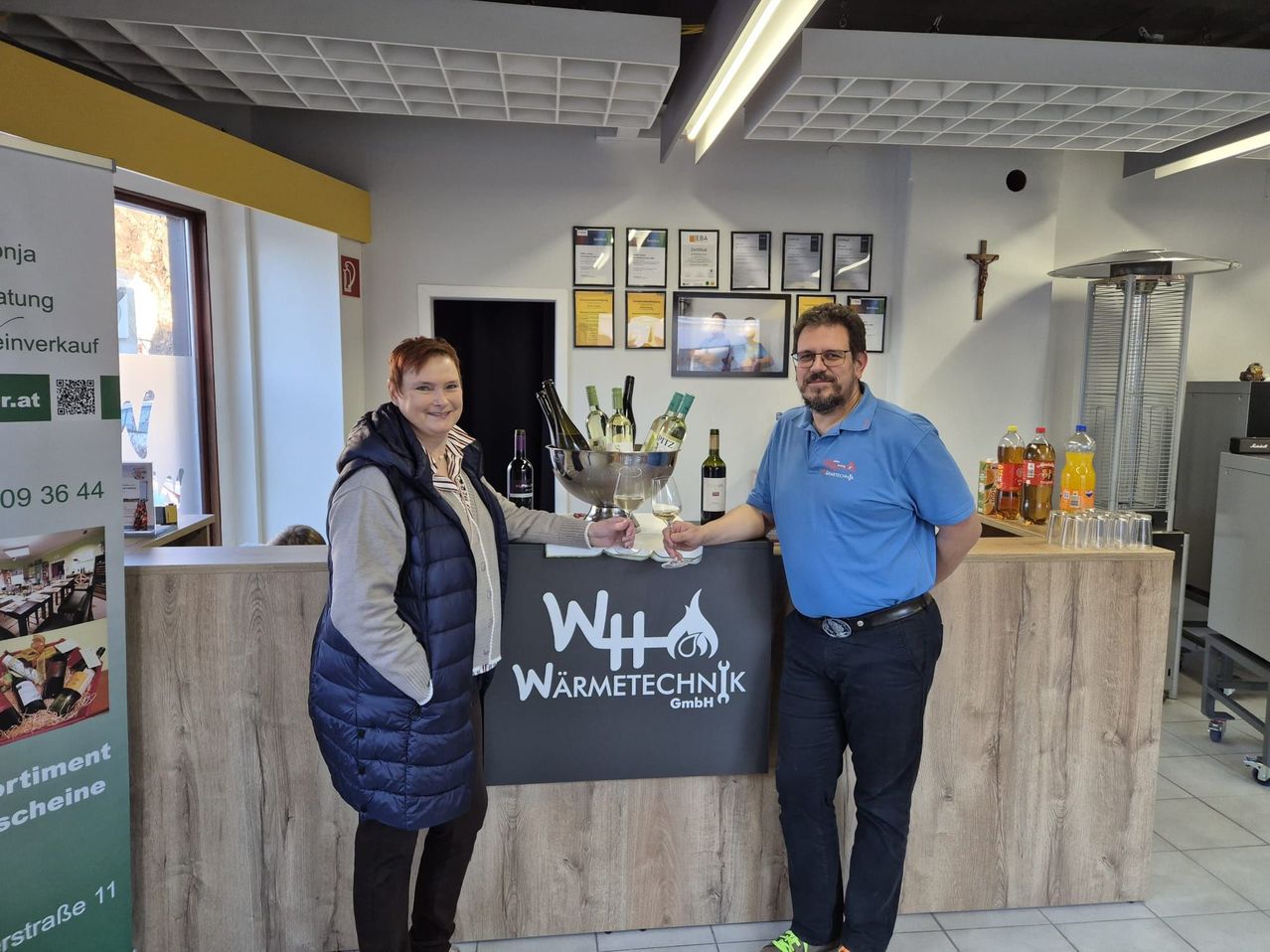 A man and a woman stand behind a desk, smiling. They hold wine glasses. Behind them is a wooden counter with a sign, bottles, and a cross.