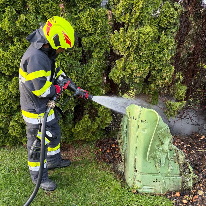 Ein Feuerwehrmann in voller Ausrüstung sprüht Wasser aus einem Schlauch auf ein ausgebranntes Fahrzeug. Die Szene spielt sich draußen ab, mit Hecken und Gras im Hintergrund.