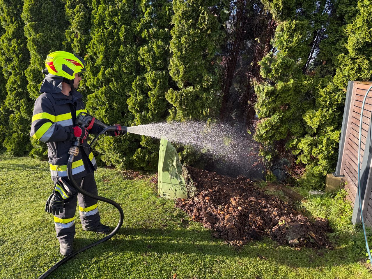 Ein Feuerwehrmann sprüht mit einem Schlauch Wasser auf einen grünen Busch, möglicherweise um ein Feuer zu löschen.