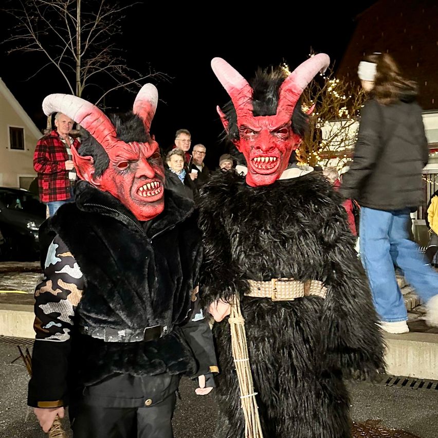 Zwei Menschen in roten Teufelskostümen posieren nachts für ein Foto. Dahinter sieht eine Menschenmenge zu. Eine Frau in einer schwarzen Jacke geht auf dem Bürgersteig. Ein Haus mit einem Baum ist im Hintergrund.
