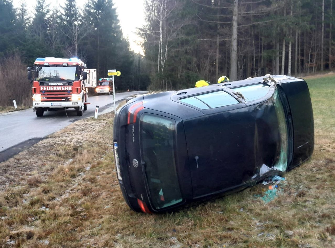 Ein umgekipptes Auto am Straßenrand, mit Rettungsfahrzeugen in der Nähe. Die Szene umfasst ein Feuerwehrauto, einen Polizeiwagen und zwei Personen in gelben Helmen. Bäume und ein Straßenschild im Hintergrund.