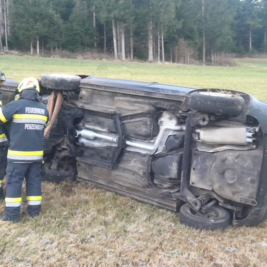 Zwei Feuerwehrleute stehen neben einem umgestürzten Auto auf einem Grasfeld, einer hält ein Werkzeug. Die Reifen des Autos sind vom Boden abgehoben.