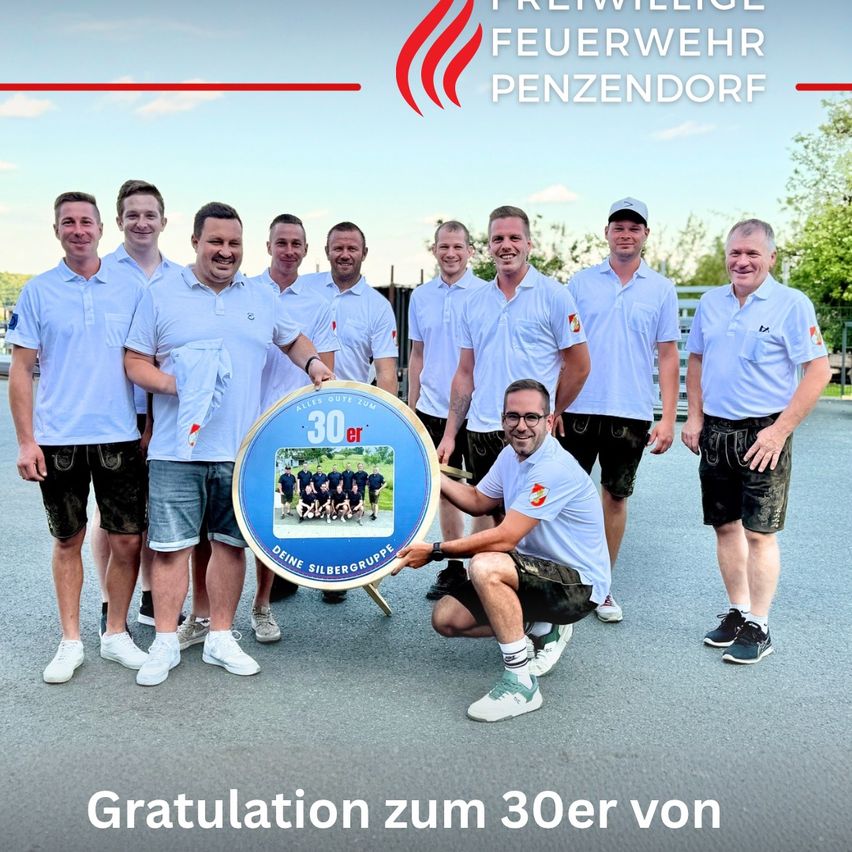 Eine Gruppe von Männern in weißen Hemden, Shorts und Turnschuhen steht mit einer großen blauen Plakette und feiert das 30-jährige Jubiläum einer Feuerwehr.