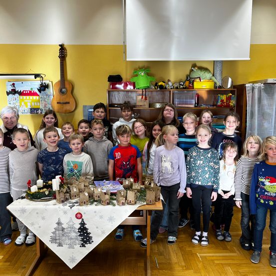 Eine Gruppe von Kindern in einem Klassenzimmer mit einem Tisch voller Lebkuchenhäuser und Kerzen, sie stehen hinter dem Tisch und lächeln in die Kamera. Eine Gitarre und verschiedene Spielzeuge sind im Hintergrund zu sehen.