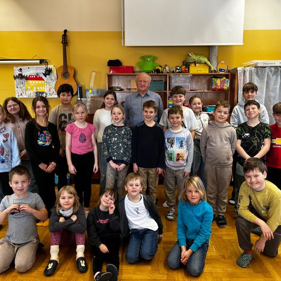 Eine Gruppe von Kindern und einem Mann in einem Klassenzimmer. Einige stehen, einige knien und alle lächeln. Das Klassenzimmer hat gelbe Wände, einen Holzboden und Regale mit verschiedenen Gegenständen.