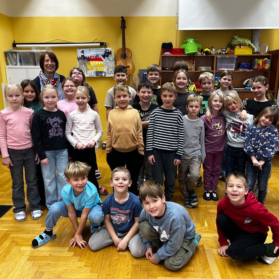 Ein Klassenzimmer voller Kinder und eine Lehrerin. Die Kinder stehen und knien, lächeln und einige schauen in die Kamera. Die Lehrerin steht hinter ihnen und lächelt. Es gibt eine gelbe Wand, Regale mit Spielzeug und eine Gitarre im Hintergrund.
