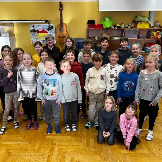 Ein Klassenzimmer voller Kinder und Erwachsener; eine Lehrerin steht in der Mitte, und eine Gitarre hängt an der Wand. Die Kinder posieren für ein Foto.
