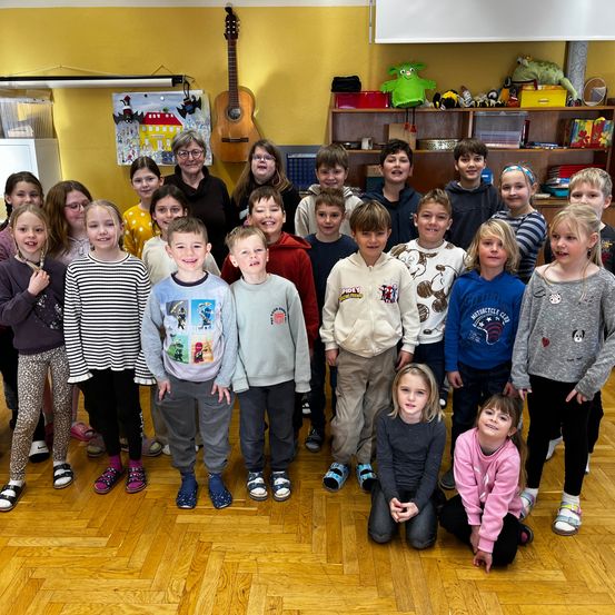 Ein Klassenzimmer voller Kinder und Erwachsener; eine Lehrerin steht in der Mitte, und eine Gitarre hängt an der Wand. Die Kinder posieren für ein Foto.