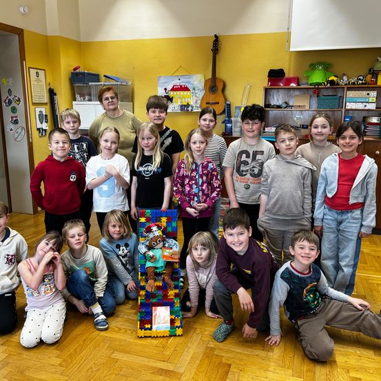 Eine Gruppe von Kindern und eine erwachsene Frau posieren für ein Foto in einem Raum. Sie sind von Spielzeug und einer Gitarre an einem Regal umgeben.