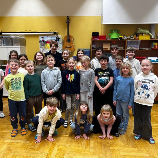 Eine Gruppe von Kindern und einer Lehrerin in einem Klassenzimmer mit einer Gitarre an der Wand. Die Kinder lächeln und posieren für ein Foto.