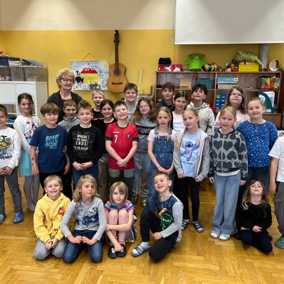Eine Gruppe von Kindern und einer erwachsenen Frau in einem Klassenzimmer, lächelnd und posierend für ein Foto. Der Raum ist mit gelben Wänden, einem Regal mit Spielzeug und einer Gitarre dekoriert.