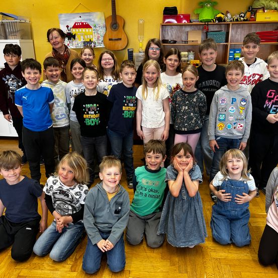 Eine Gruppe von Kindern im Klassenzimmer, einige knien, andere stehen und lächeln in die Kamera. Eine Frau steht hinter ihnen und eine Gitarre hängt an der Wand.