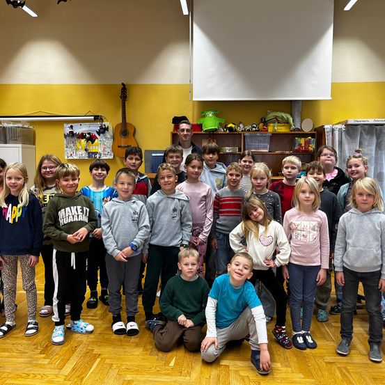Eine Gruppe von Kindern und einigen Erwachsenen stehen und sitzen in einem Raum. Die Kinder tragen Freizeitkleidung, einige haben Schuhe an, andere sind barfuß. Eine Gitarre und eine Tafel sind im Hintergrund zu sehen.
