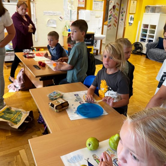Ein Klassenzimmer mit Kindern, die essen, einer Frau im Hintergrund und einem Ventilator. Ein Kind schaut auf Früchte auf einem Schreibtisch.