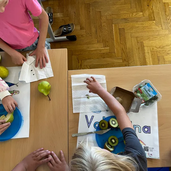 Kinder sitzen an einem Tisch, jeder mit einer Birne und einem Teller. Ein Kind hat Kiwi auf seinem Teller und sie halten alle Servietten. Der Boden ist aus Holz.