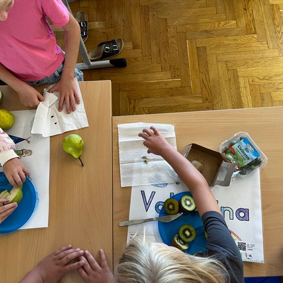 Kinder sitzen an einem Tisch, jeder mit einer Birne und einem Teller. Ein Kind hat Kiwi auf seinem Teller und sie halten alle Servietten. Der Boden ist aus Holz.