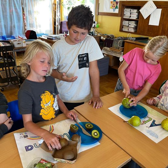 Drei Kinder in einem Klassenzimmer schneiden Kiwis und Birnen auf blauen Tellern. Ein Junge trägt ein graues Shirt mit einem Comic-Charakter. Ein weiterer Junge im weißen Shirt steht hinter ihm. Ein Mädchen im rosa Shirt hält eine Birne.