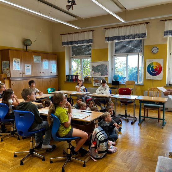 Ein Klassenzimmer mit Kindern an Schreibtischen und Stühlen, einer Lehrerin vorne, einem Globus und einer Uhr an der Wand.