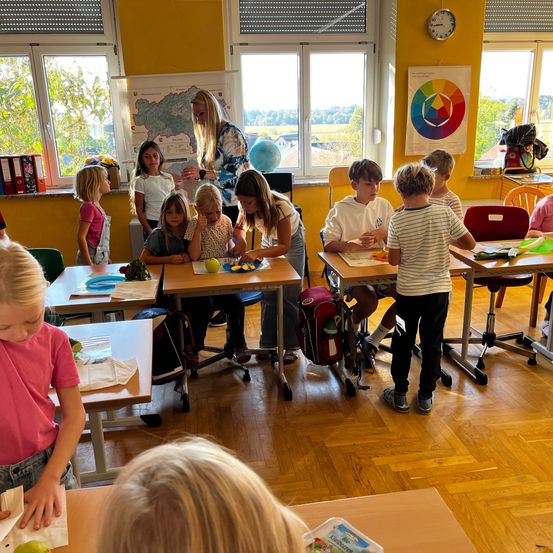 Kinder sitzen in einem Klassenzimmer an Tischen und bereiten Äpfel für einen Snack vor. Eine Lehrerin steht in der Nähe und hilft. Gelbe Wände, große Fenster und ein Globus sind sichtbar.