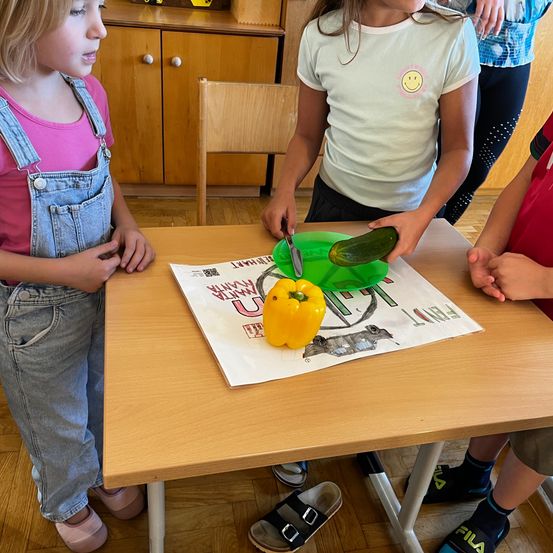 Drei junge Kinder stehen um einen Tisch herum. Eines hält einen grünen Teller mit einer Gurke und einer gelben Paprika. Die anderen beiden beobachten.