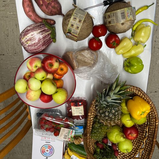 Ein Tisch zeigt eine Auswahl an Obst und Gemüse. Ein Korb mit Äpfeln und eine Schale mit Ananas, Paprika und anderem Gemüse sind zu sehen.
