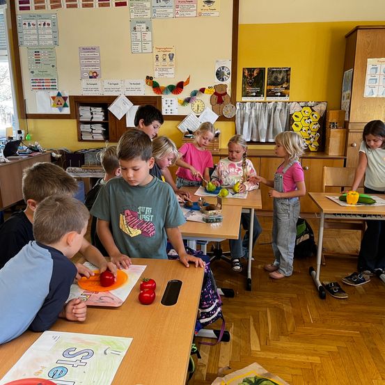 Kinder in einem Klassenzimmer sind mit einer Essenszubereitung beschäftigt, wobei einige Gemüse schneiden, während andere zusehen. Der Raum hat Holzböden und ist mit pädagogischen Postern geschmückt.