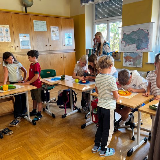 Kinder versammeln sich in einer Klasse um Tische herum und beschäftigen sich unter Aufsicht einer erwachsenen Frau mit dem Schneiden von Gemüse. Jedes Kind hat ein Messer und einen Teller mit Gemüse.