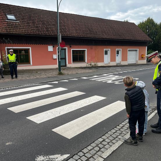 Ein Polizist steht in der Nähe einer Fußgängerampel und leitet zwei Kinder. Ein rotes Gebäude mit Glasfenstern steht dahinter. Der Himmel ist bewölkt.