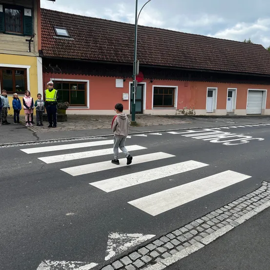 Ein Junge läuft über einen Zebrastreifen, bewacht von einem Verkehrspolizisten und Kindern. Ein Gebäude mit braunen Ziegeln und eine Straßenlaterne stehen im Hintergrund.
