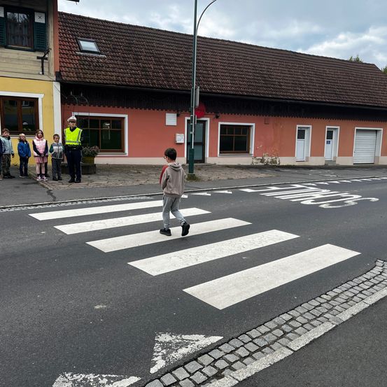 Ein Junge läuft über einen Zebrastreifen, bewacht von einem Verkehrspolizisten und Kindern. Ein Gebäude mit braunen Ziegeln und eine Straßenlaterne stehen im Hintergrund.