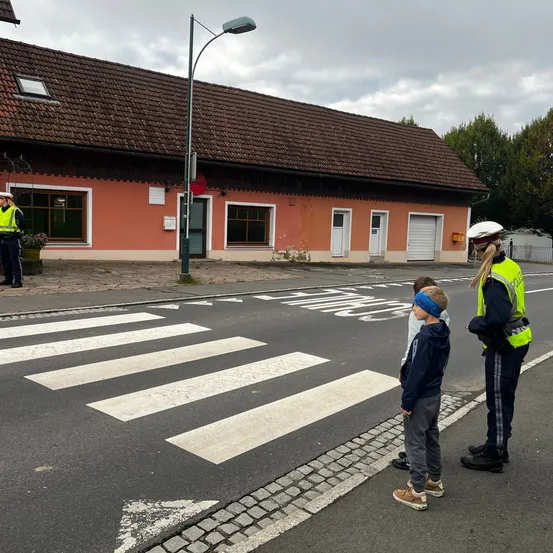 Ein Verkehrspolizist lehrt zwei Kinder, wie man sicher die Straße auf einem Zebrastreifen überquert. Ein Gebäude mit braunem Dach und ein Polizist sind im Hintergrund.