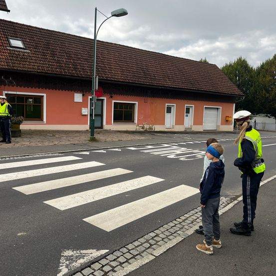 Ein Verkehrspolizist lehrt zwei Kinder, wie man sicher die Straße auf einem Zebrastreifen überquert. Ein Gebäude mit braunem Dach und ein Polizist sind im Hintergrund.