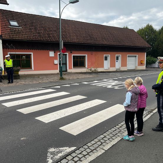 Zwei junge Mädchen stehen auf einem Zebrastreifen, überwacht von einem Verkehrspolizisten. Hinter ihnen steht ein Haus mit einem braunen Dach, ein Mann in einem gelben Westen steht neben dem Gebäude.