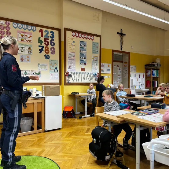 Eine Polizistin steht in einem Klassenzimmer und spricht mit aufmerksamen Kindern an ihren Schreibtischen. Der Raum ist mit pädagogischen Postern und einem Kreuz an der Wand ausgestattet.