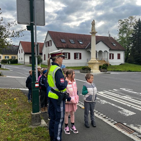 Eine Polizistin weist eine Gruppe von Kindern auf dem Bürgersteig an. Sie befinden sich in der Nähe eines Denkmals mit einer Statue. Die Kinder tragen Winterkleidung, und die Polizistin ist in Uniform.